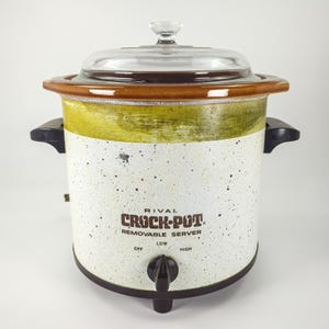 May include: A vintage Rival Crock-Pot removable server with a white speckled body, a green band near the top, and a brown rim. It features a clear glass lid, two black side handles, and a black control knob with Off, Low, and High settings.