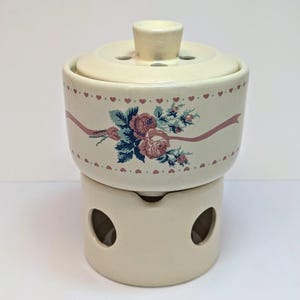 May include: A cream-coloured ceramic warmer featuring a removable lid with ventilation holes. The cylindrical body is decorated with a horizontal pink ribbon, a floral bouquet of pink roses and green leaves, and a border of small pink hearts.