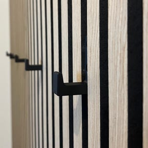 Könnte beinhalten: Nahaufnahme einer Wand mit schwarzen und weißen vertikalen Streifen. Drei schwarze Metallhaken sind an der Wand befestigt.
