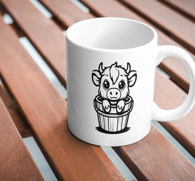 Puede incluir: Taza de cer&aacute;mica blanca con una ilustraci&oacute;n en blanco y negro de una vaca de dibujos animados en un cubo. La vaca tiene ojos grandes y mira al espectador.