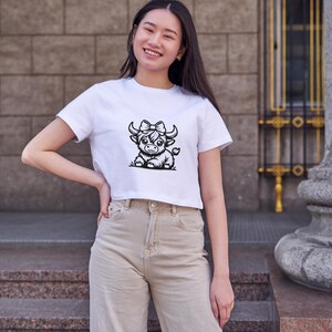 Puede incluir: Una camiseta blanca recortada con una ilustraci&oacute;n en blanco y negro de una vaca de las Tierras Altas con un lazo. La vaca mira hacia la derecha.
