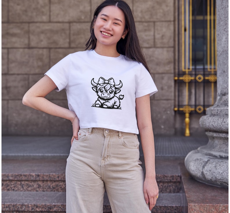 Puede incluir: Una camiseta blanca recortada con una ilustraci&oacute;n en blanco y negro de una vaca de las Tierras Altas con un lazo. La vaca mira hacia la derecha.