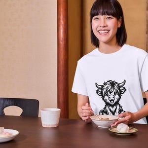 Puede incluir: Una mujer lleva una camiseta blanca con una ilustraci&oacute;n en blanco y negro de una vaca de las Tierras Altas en la parte delantera. Est&aacute; sentada en una mesa con un cuenco de comida y una taza.
