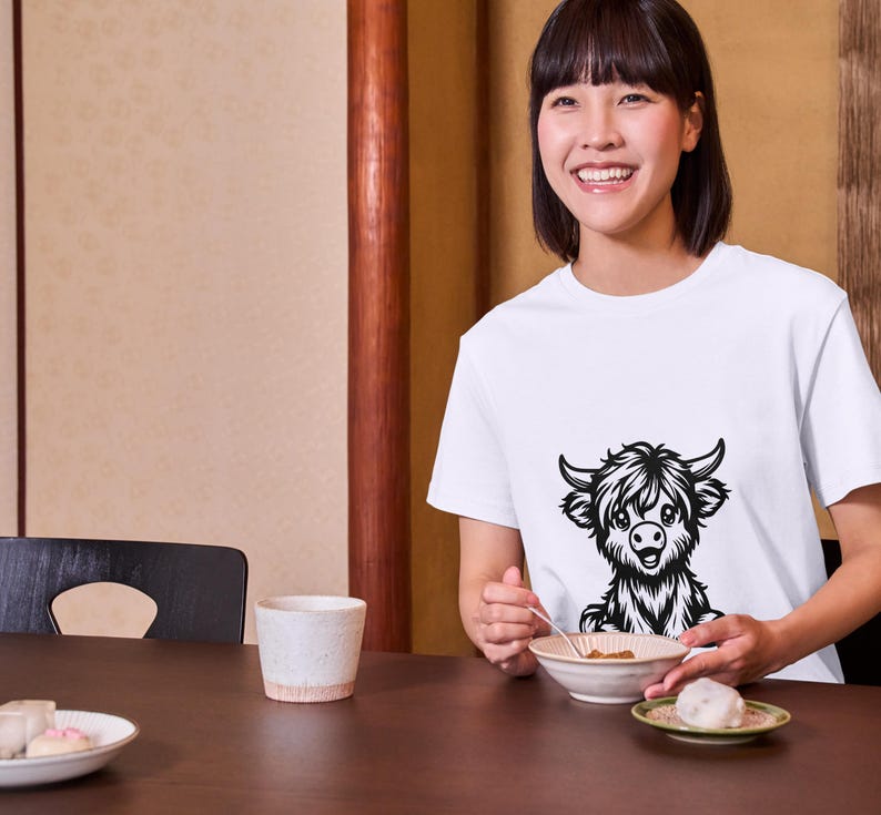 Puede incluir: Una mujer lleva una camiseta blanca con una ilustraci&oacute;n en blanco y negro de una vaca de las Tierras Altas en la parte delantera. Est&aacute; sentada en una mesa con un cuenco de comida y una taza.