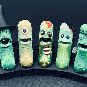 May include: A collection of five green, cucumber-shaped figurines with various monster-themed designs. One wears a black witch hat, while others feature zombie and Frankenstein-inspired details. They are displayed on a black, curved stand.