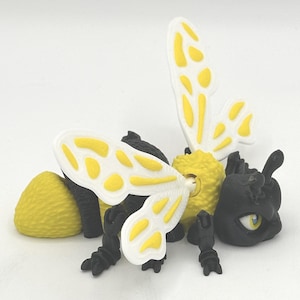 Puede incluir: Un juguete de abeja negro y amarillo con alas blancas. La abeja tiene un cuerpo segmentado y ojos grandes y negros. Las alas tienen manchas amarillas.