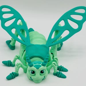 Puede incluir: Un insecto de juguete verde con alas grandes y una cara amigable. El insecto tiene un cuerpo segmentado y patas largas.