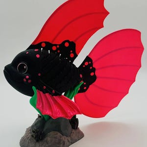 Peut inclure: Sculpture de poisson noir fantaisiste avec de grandes nageoires et des accents rouges vifs. Le poisson a des taches roses et est posé sur un socle en roche grise avec des détails verts et roses. Objet décoratif.