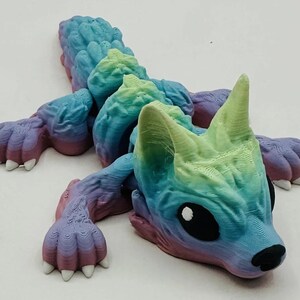 Puede incluir: Un juguete articulado y colorido con forma de criatura en tonos azules, verdes, morados y rosas. El juguete tiene una cabeza de lobo con grandes ojos negros y una nariz negra. El cuerpo está segmentado y las patas tienen garras blancas.