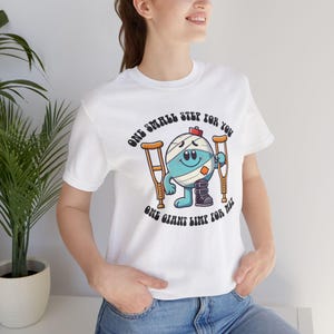 Peut inclure: T-shirt blanc avec un personnage de dessin animé portant des bandages et tenant des béquilles. Le texte sur le t-shirt se lit : « UN PETIT PAS POUR VOUS, UN GRAND MEMBRE POUR MOI ».