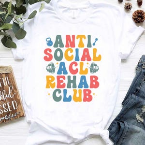 May include: White t-shirt featuring the text "ANTI SOCIAL ACL REHAB CLUB" in a retro font, with dumbbell and kettlebell illustrations. The design uses a palette of bright colours. The t-shirt is made of a soft material.