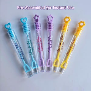 May include: Six clear bubble wands with colored handles and tops. The handles are blue, purple, and yellow. Each wand has the text "Letis & John" and the date "08.16.2026". The tops are shaped like hearts, stars, and flowers.
