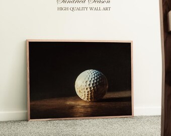 Op de afbeelding: Een ingelijste wandkunstprint met een golfbal. De golfbal is wit met putjes en wordt verlicht door een enkele lichtbron. De print bevat de tekst "Sundried Season" en "HIGH QUALITY WALL ART".