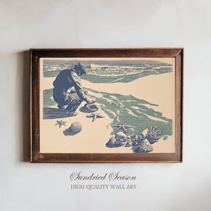 Può includere: Una stampa incorniciata raffigurante una scena di spiaggia con una persona che raccoglie conchiglie. L'opera d'arte è nei toni del blu e del beige, con il testo "Sundried Season" e "HIGH QUALITY WALL ART" in basso.