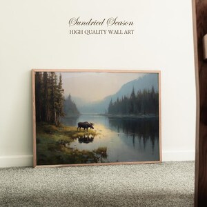 Op de afbeelding: Een ingelijste wandkunstprint met een serene landschap met een eland bij een meer. Het kunstwerk heeft een natuurlijke houten lijst en de tekst "Sundried Season HIGH QUALITY WALL ART" bovenaan.