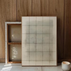 May include: Abstract artwork with a grid pattern in black lines on a cream background, framed in natural wood. A smaller, white-framed window-like structure is to the left. A lit candle in a white holder is on the right.