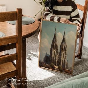 May include: A framed print of a cathedral with two tall spires, set against a blue sky with clouds. The print has a wooden frame and is held by a person. The text "Sundried Seas" and "HIGH QUALITY WALL ART" is visible at the bottom.