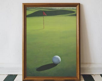 Op de afbeelding: Een ingelijste prent van een golfbaan. Het kunstwerk toont een golfbal op een green, met een rode vlag in de verte. De lijst is bruin van kleur, en de achtergrond is een witte muur en een zwart-wit gestreepte vloer.