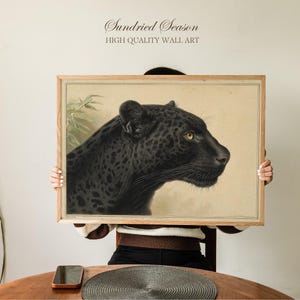 Op de afbeelding: Een ingelijste print van een zwarte jaguar met gele ogen, tegen een beige achtergrond. Het kunstwerk zit in een houten lijst en bevat de tekst "Sundried Season HIGH QUALITY WALL ART".