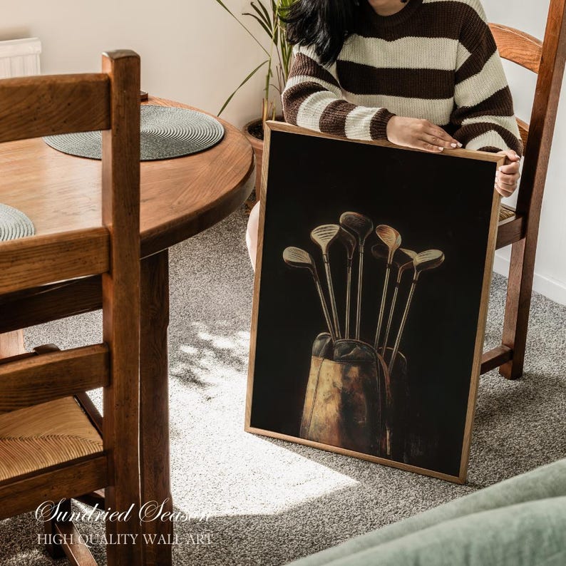 Op de afbeelding: Een ingelijste print met golfclubs in een tas tegen een zwarte achtergrond. De print heeft een houten lijst en wordt door een persoon vastgehouden. De tekst "Sundried Seas" en "HIGH QUALITY WALL ART" staat onderaan.