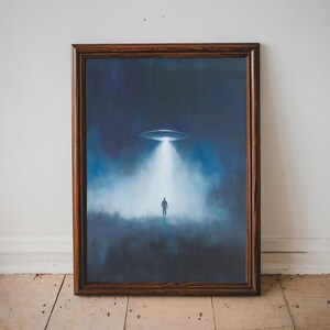 May include: Framed artwork depicting a nighttime scene with a UFO shining a bright light on a silhouetted figure. The artwork has a dark blue and white color palette, with a brown wooden frame. The artwork is displayed against a white wall and wooden floor.