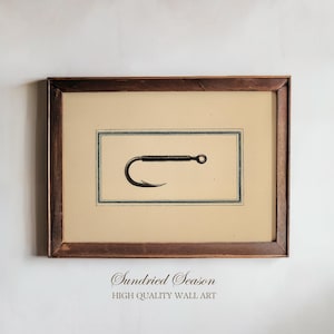 Puede incluir: Ilustración enmarcada de un anzuelo de pesca. La obra de arte está en un marco rectangular de madera marrón oscuro. El anzuelo es negro, sobre un fondo crema con un borde verde. El texto "Sundried Season" y "HIGH QUALITY WALL ART" está en la parte inferior de la impresión.