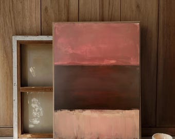 May include: Abstract painting in a wooden frame with horizontal bands of color: dusty rose, deep brown, and pale pink. The artwork leans against a wooden wall next to a white-framed window and a lit candle in a textured ceramic holder.