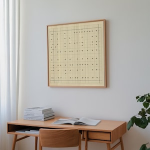 May include: A framed artwork featuring a grid-like pattern of black dots on a cream-colored background. The artwork is displayed on a white wall above a wooden desk with books and an open notebook. The frame is a light brown color.