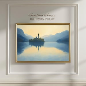 Può includere: Un dipinto di paesaggio incorniciato con una serena scena lacustre con una piccola isola e una chiesa. L'opera d'arte è in una cornice dorata ed è montata su una parete bianca. Il testo "Sundried Season HIGH QUALITY WALL ART" è sopra.