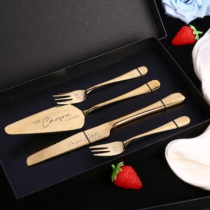 Peut inclure: Un ensemble de service à gâteau doré, comprenant un couteau à gâteau, une pelle et deux fourchettes, présenté dans un coffret noir. Le couteau et la pelle sont gravés des noms "Carson & Hale" et de la date "12.05.2026".