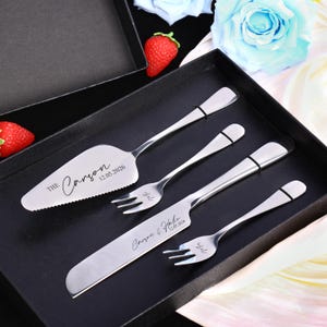 Può includere: Un set da portata per torte argentato con una paletta per torte, un coltello e due forchette, presentato in una scatola nera. La paletta e il coltello sono incisi con nomi e una data. Un regalo di nozze o anniversario.