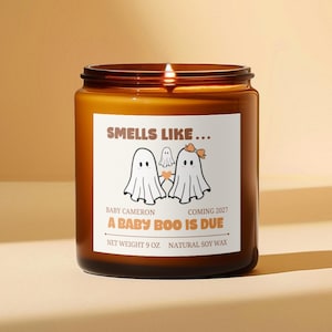 May include: A brown glass jar candle with a white label featuring the text "SMELLS LIKE... A BABY BOO IS DUE", "BABY CAMERON", and "COMING 2027". The label displays three ghost illustrations, one with a bow, and a small heart. Net weight 9 oz.