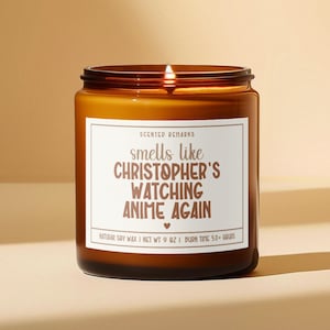 May include: A brown glass candle with a white label that reads "SCENTED REMARKS smells like CHRISTOPHER'S WATCHING ANIME AGAIN" with a small heart symbol. The candle is lit and the wax is melting. The label also says "NATURAL SOY WAX | NET WT 9 OZ | BURN TIME 50+ HOURS".