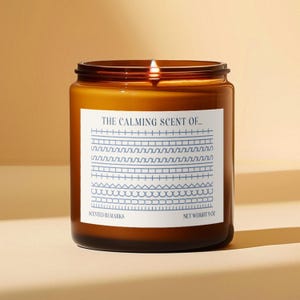 May include: A lit, amber-colored scented candle in a glass jar. The label reads "THE CALMING SCENT OF..." with blue geometric designs. The net weight is 9 oz.