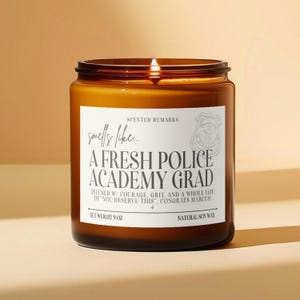 May include: A brown glass candle with a white label that reads "smells like... A FRESH POLICE ACADEMY GRAD INFUSED W/ COURAGE, GRIT, AND A WHOLE LOT OF 'YOU DESERVE THIS', CONGRATS MARCUS!"  The label also includes the text "SCENTED REMARKS", "NET WEIGHT 9 OZ", and "NATURAL SOY WAX".