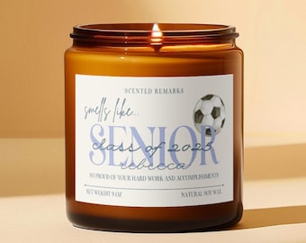 May include: A brown glass jar candle with a white label featuring a soccer ball graphic and text that reads Scented Remarks, smells like... SENIOR, class of 2025, rebecca, so proud of your hard work and accomplishments, net weight 9 oz, natural soy wax.