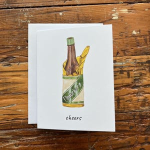 Puede incluir: Tarjeta de felicitación blanca con una ilustración de una botella de Underberg en una lata, con un gesto de la mano amarilla. La palabra "cheers" está escrita debajo. La tarjeta está sobre un fondo de madera.