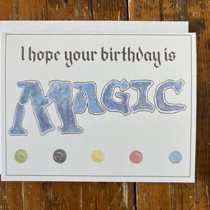 May include: A birthday card with the words "I hope your birthday is MAGIC" in a whimsical font. The word "MAGIC" is in blue watercolor lettering. The card has a white border and colorful dots.