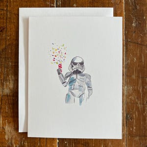 May include: A white greeting card with a watercolor illustration of a Stormtrooper holding a party popper. The party popper is red and white, with colorful confetti bursting from it. The card is blank inside.