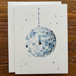 May include: A white greeting card with a watercolor illustration of a disco ball in shades of blue. The disco ball is hanging from a thin line, with small blue accents around it. The card is paired with a white envelope.