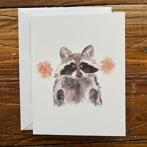May include: A watercolor greeting card featuring a raccoon with a black mask and fluffy gray fur. The raccoon is centered on the white card, with two pink and yellow floral accents on either side. A white envelope is behind the card.
