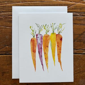 Puede incluir: Pintura a la acuarela de cinco zanahorias en tonos naranja, morado y amarillo, cada una con hojas verdes. La obra de arte está en una tarjeta blanca, con un sobre blanco en blanco detrás. El fondo es una superficie de madera.