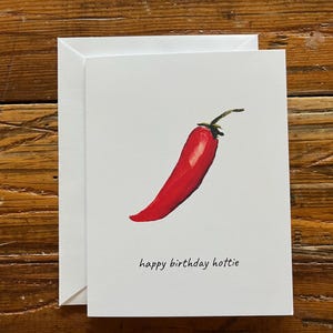 Può includere: Un biglietto di auguri bianco con un'illustrazione di peperoncino rosso e la scritta "happy birthday hottie". Il biglietto è accompagnato da una busta bianca. L'immagine è ambientata su uno sfondo di legno.