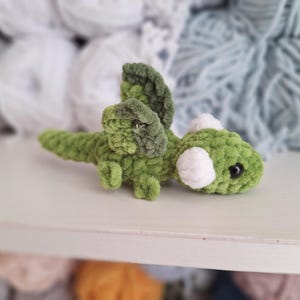 May include: A handmade green and white crochet dragon toy. The dragon has a long tail, wings, and white accents. The toy is made of soft, textured yarn. The dragon is lying on a white surface.