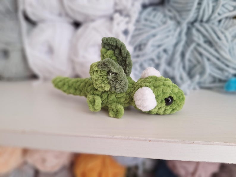 May include: A handmade green and white crochet dragon toy. The dragon has a long tail, wings, and white accents. The toy is made of soft, textured yarn. The dragon is lying on a white surface.