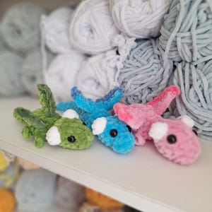 May include: Four small, crocheted dragon toys in shades of green, blue, and pink with white accents and black eyes. The toys are displayed on a white shelf, with balls of yarn in the background.