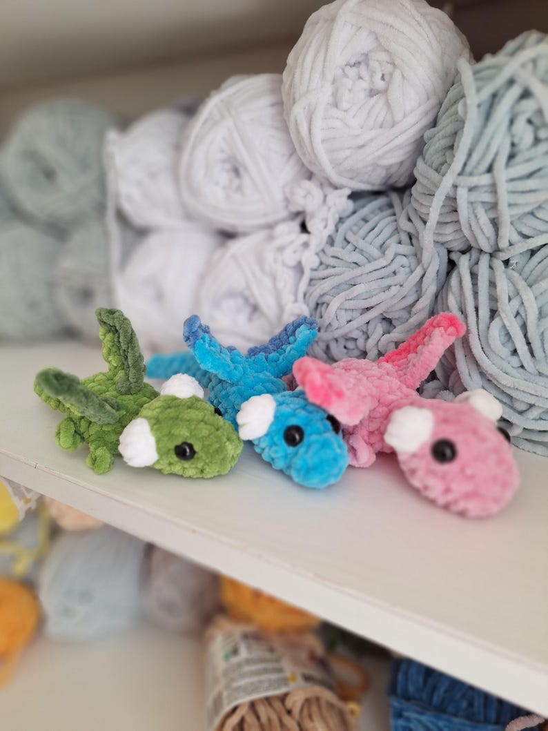 May include: Four small, crocheted dragon toys in shades of green, blue, and pink with white accents and black eyes. The toys are displayed on a white shelf, with balls of yarn in the background.