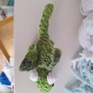 May include: A handmade green crochet alligator toy with white eyes. The alligator is made of a soft, textured yarn. The toy is lying on a white surface. Crochet toys are a popular item for children.