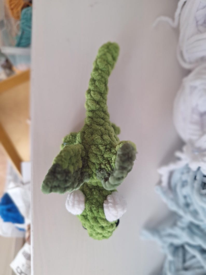 May include: A handmade green crochet alligator toy with white eyes. The alligator is made of a soft, textured yarn. The toy is lying on a white surface. Crochet toys are a popular item for children.