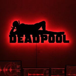 May include: A red-lit wall decoration featuring the silhouette of Deadpool in a relaxed pose, with the word "DEADPOOL" beneath. The room is illuminated in red, with computer monitors and keyboards visible below.